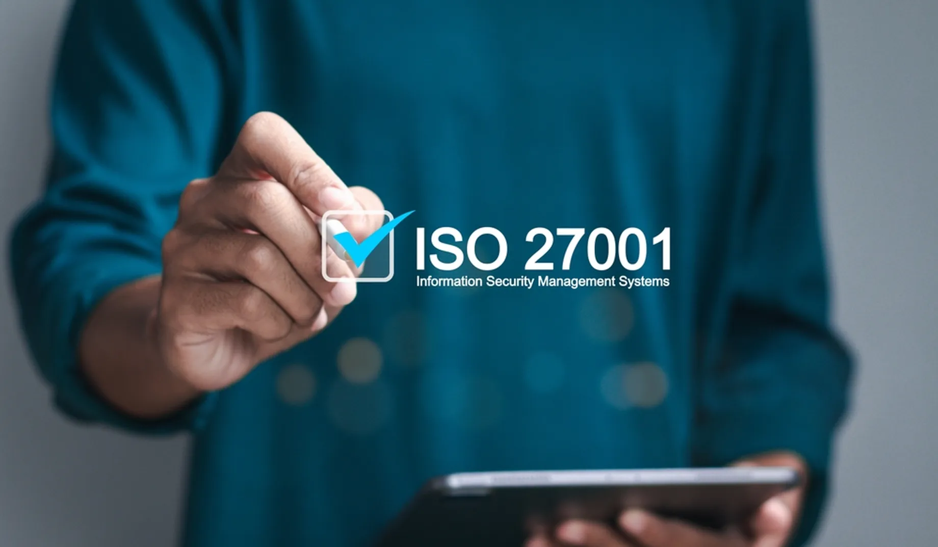 Person in blue shirt holding tablet and drawing a check mark next to text “ISO 27001 Information Security Management Systems.”
