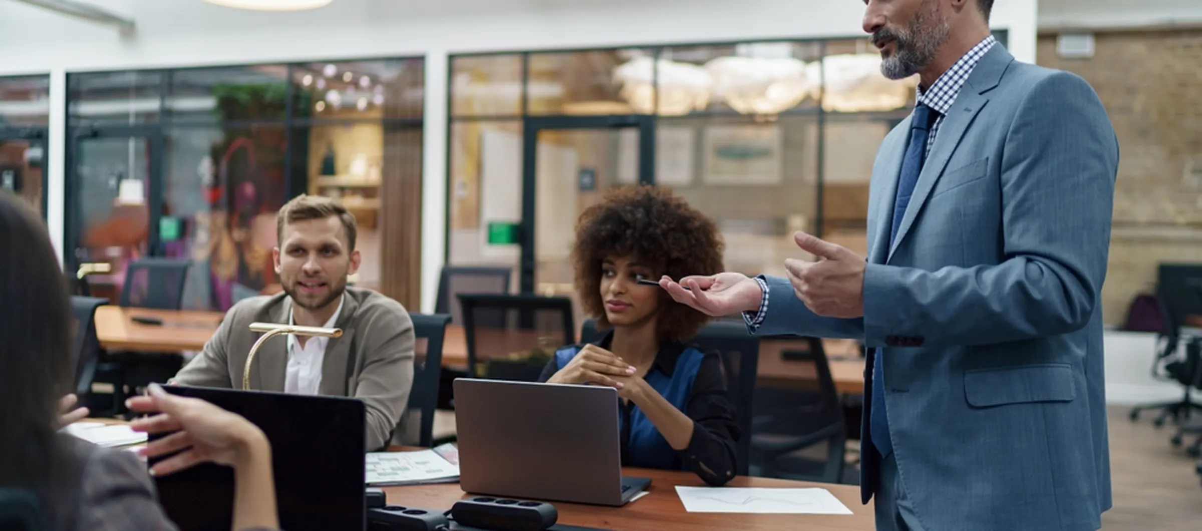 Business team in a modern office, with a man in a suit leading a meeting while colleagues listen and work on laptops around a table.