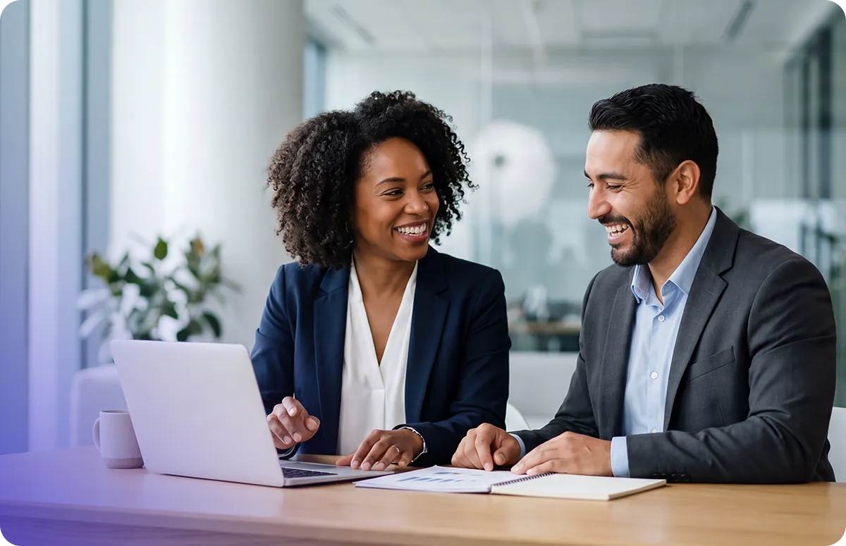 Two business professionals smiling and discussing work at a laptop in a modern office meeting.