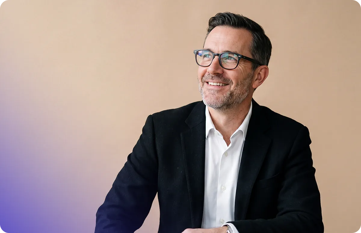 Smiling man in glasses and a blazer looks off to the side against a beige and purple gradient background.
