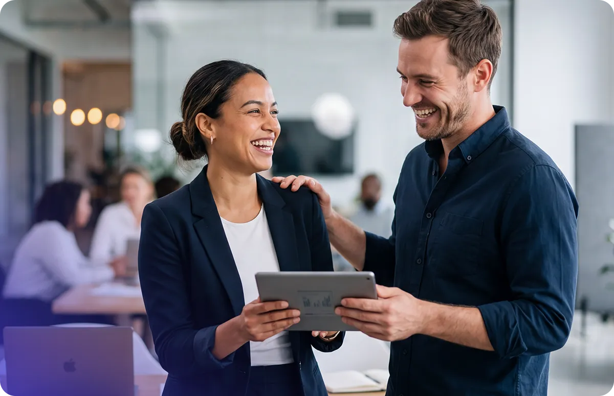 Two coworkers smiling and looking at a tablet together in a modern open office setting.