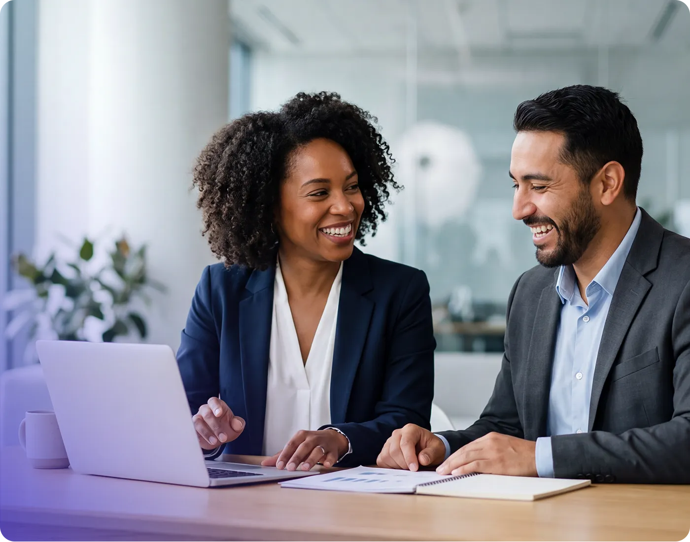 Two business professionals in suits smile and discuss documents at a desk with a laptop in a bright modern office.