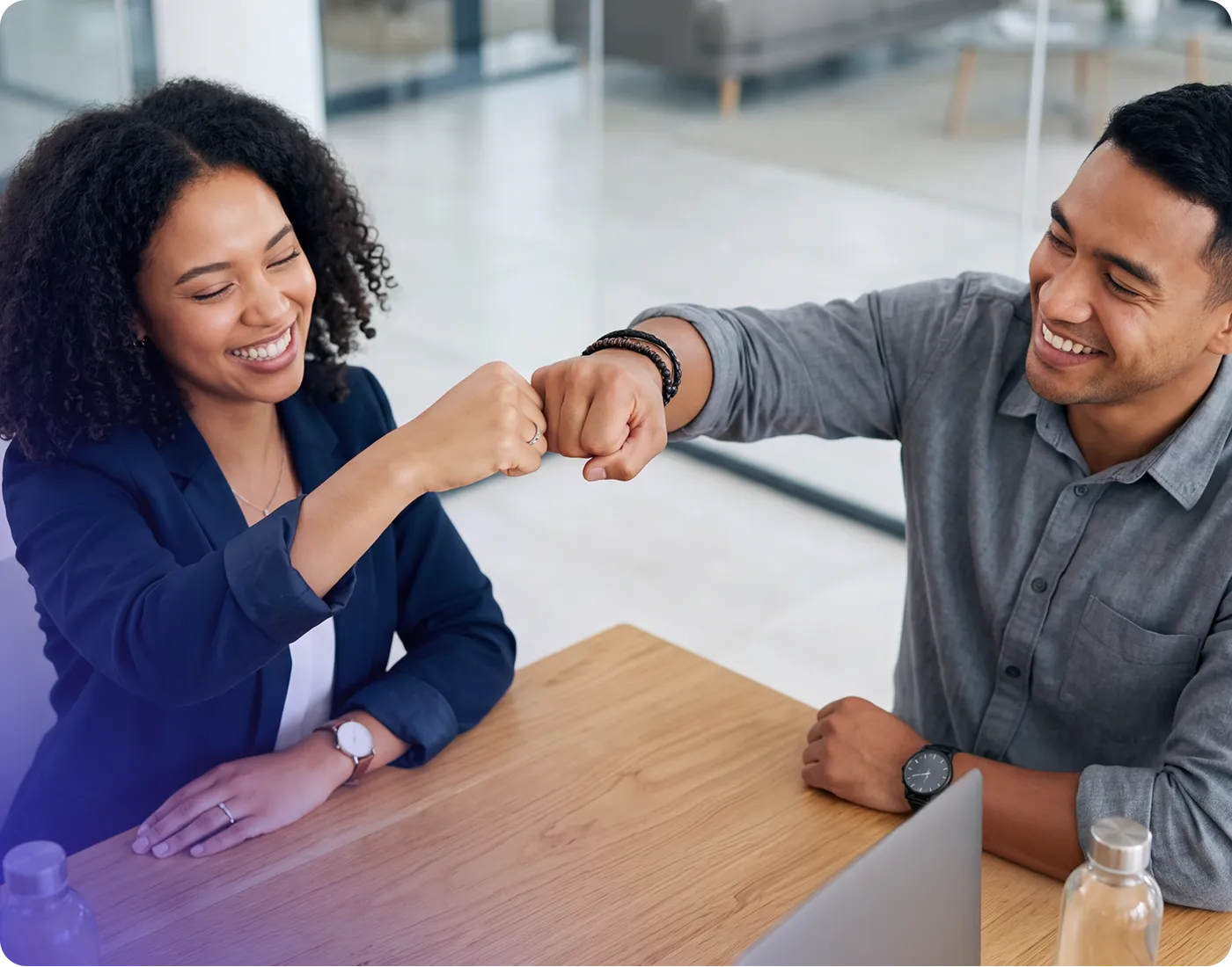 Two smiling coworkers sit at a desk, leaning forward to give each other a celebratory fist bump in a modern office.