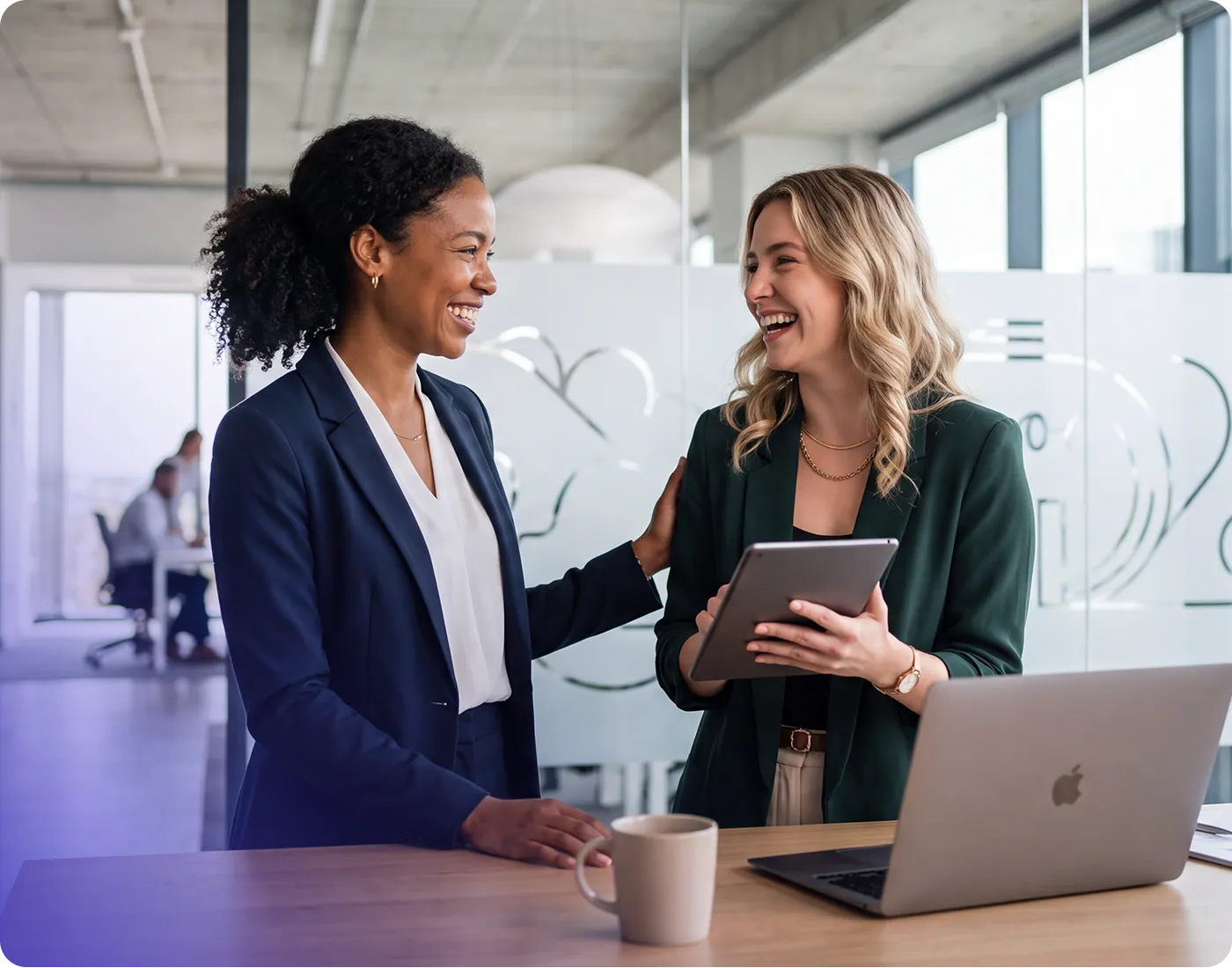 Two businesswomen standing by a desk, smiling and talking while one holds a tablet in a modern glass-walled office.