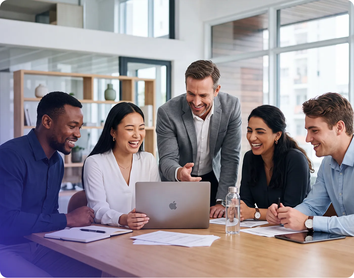 Diverse group of five colleagues laughing and collaborating around a laptop in a bright modern office meeting room.